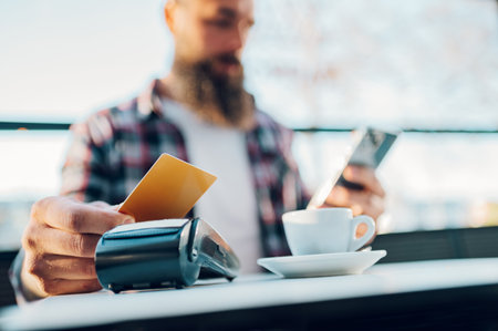 Bearded man in casual outfit using credit and paying on a payment terminal while sitting at cafe...