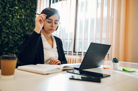 Young caucasian businesswoman smiling while sitting at her desk in a bright modern office working on a laptop with coworkers in the background. Startup business in development.の写真素材