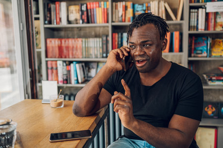 Cheerful african american man talking on his smartphone while sitting in a cafe. Messaging online and checking news feed on social networks during coffee break in modern restaurant interior.の写真素材
