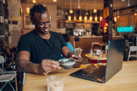 Cheerful african american businessman having coffee and doing his work in cafe while using a laptop. Modern lifestyle, connection, business, freelance work concept.の写真素材