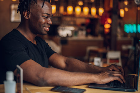 Smile african american business man sitting in cafe holding a cup of coffee and using laptop. Creates new content for his blog, enjoys the work of a freelancer.の写真素材