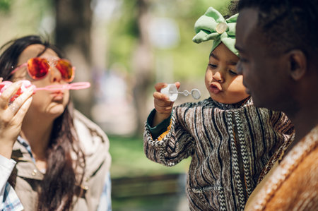 Diverse family blowing bubbles together in the park. Multiracial parents and their baby girl having...