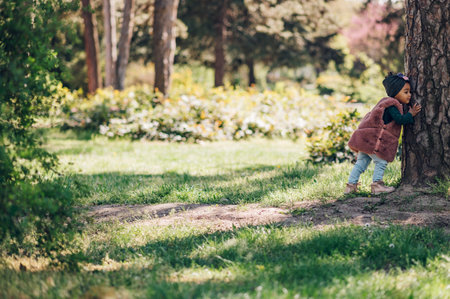 Happy little african american baby girl walking in a park. Beautiful black little girl laughing and playing outside. Hugging a tree. Little nature lover.の写真素材