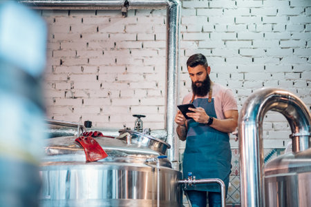 Portrait of brewery worker checking fermentation process and using application on a tablet for a...