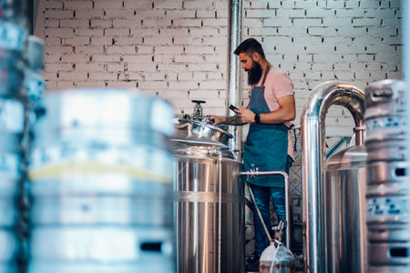 Smiling bearded handsome man in apron using tablet and checking data on large metal tank with craft beer in a brewery. Craft beverage production. Fermentation process in plant.の写真素材