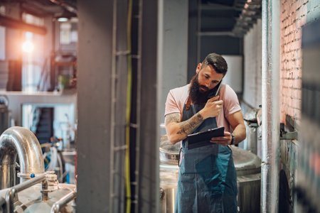 Bearded caucasian man with clipboard taking notes and using a smartphone while working at craft brewery. Manufacture, business and people concept.の写真素材