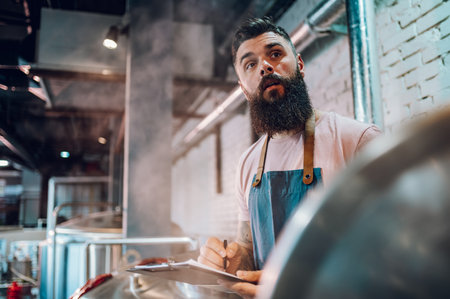 Attractive man worker wearing apron and making notes while standing near metal kettle in a beer...