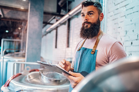 Bearded man worker in a brewery using brewing equipment at factory and writing results in a clipboard. Control of plant, collecting data. Inspecting industrial equipment and making notes.の写真素材