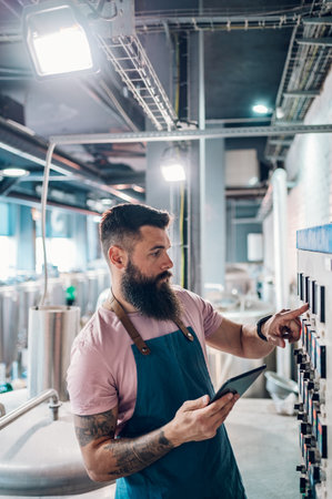Bearded man worker in a brewery wearing an apron and using brewing equipment at factory. Control of plant, collecting data. Inspecting industrial equipment and making notes.の写真素材