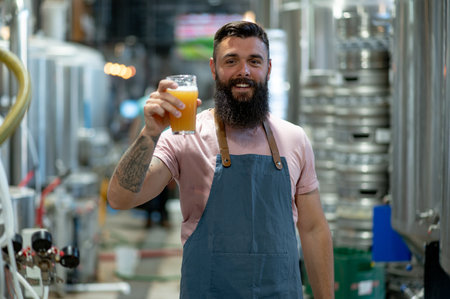 Bearded man worker examining beer glass while working at the brewery. Brewer working in his craft brewery. Wearing an apron. Focus on a man face.の写真素材