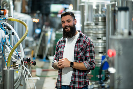 Young bearded man standing by beer filling machine and posing. Brewery worker with industrial equipment. Looking into the camera. Smiling and wearing a checkered shirt.の写真素材