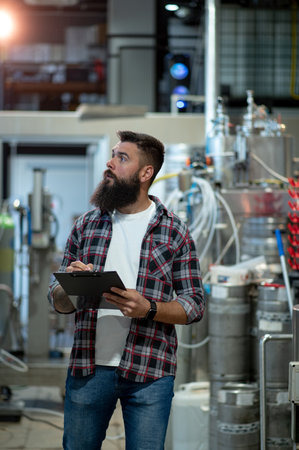 Attractive man worker wearing checkered shirt and making notes while standing near metal kettle in a beer factory interior. Beer production process, brewing at brewery. Checking beer making process.の写真素材