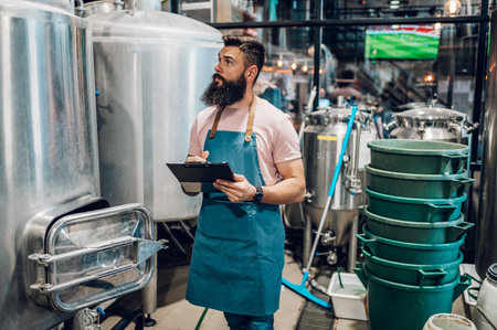 Man worker wearing apron and making notes while standing near metal kettle in a beer factory...