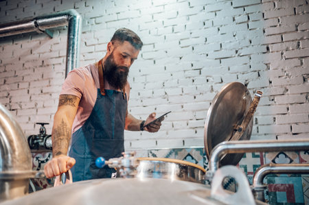 Bearded handsome man in apron using tablet and checking data on large metal tank with craft beer in a brewery. Craft beverage production. Fermentation process in plant.の写真素材