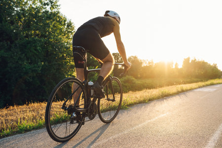 Rear view of a sporty man wearing active wear and helmet riding a black bike in nature. Concept of people, workout and favorite hobby. Sunset in the background. Copy space.の写真素材