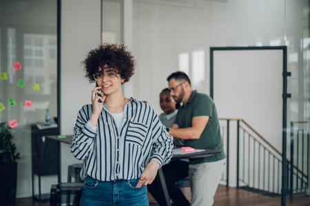 Portrait of a pretty young hispanic businesswoman with afro hair smiling and using a smartphone during a meeting in an office with her colleagues in background. Business woman team leader concept.の写真素材