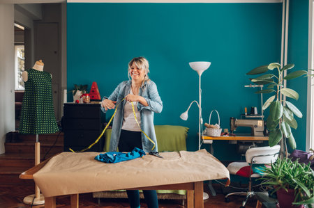 Concentrated middle aged woman tailor making pattern dress detail on sketch lines for sewing machine. Workplace of a dressmaker. Handmade concept. Using tailor's meter.の写真素材