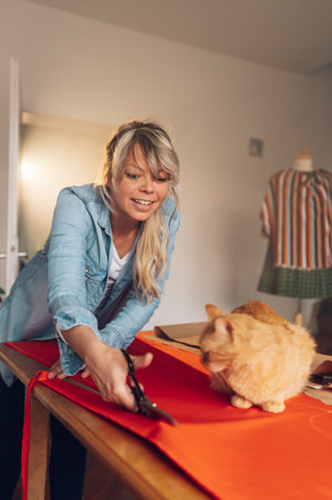 Beautiful creative woman holding scissors and cutting textile on the table at home while her pet cat is making her company. Working from home with a pet cat. Life with pets.の写真素材