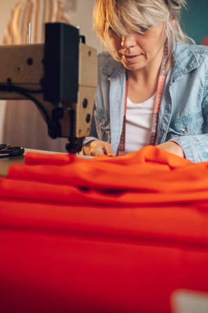 Beautiful middle aged tailor woman sewing on a sewing machine while sitting at her working place at home. Fashion atelier, tailoring, handmade clothes concept. Slow Fashion.の写真素材