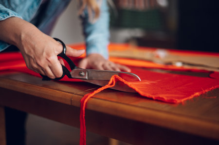 Creative middle aged woman tailor tailoring while using scissors and cutting the fabric. Woman seamstress working from home and tailoring beautiful handmade dresses. Focus on tailor hands and scissorsの写真素材
