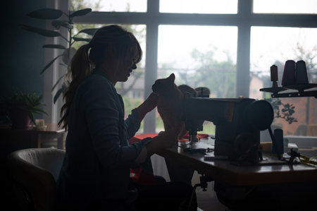 Silhouette of a blonde middle aged tailor woman sews clothes on sewing machine while working at home and petting her cat. Dressmaker working on the sewing machine. Fashion designer concept.の写真素材