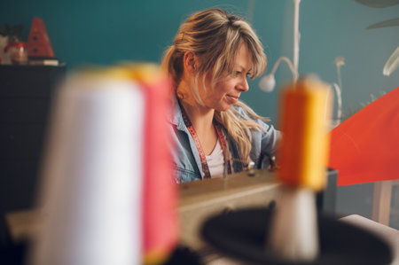 Happy middle aged dressmaker woman sitting and sewing on a sewing machine a in studio. Tailor woman...