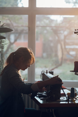 Blonde middle aged tailor woman sews clothes on sewing machine while working at home in a moody atmosphere. Dressmaker working on the sewing machine. Fashion designer concept. Silhouette of a woman.の写真素材