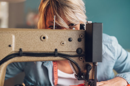 Beautiful middle aged tailor woman sewing on a sewing machine while sitting at her working place at...
