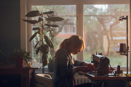 Middle aged female fashion designer pushes fabric material through a sewing machine and making a new handmade suit while working at home. Hobby sewing as a small business concept. Moody atmosphere.の写真素材