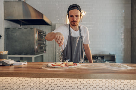 Man pizza master prepares pizza according to customer requests. Process of placing ingredients on a pizza. Pizza place and tasty food concept. Focus on a man sprinkling cheese.の写真素材