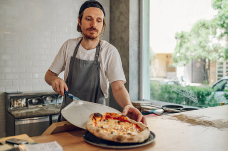 Close-up portrait a cheerful chef working in a pizza place and placing the fresh baked pizza on a wooden counter. Smiling chef wearing apron uniform and posing.の写真素材