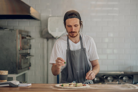 Man worker in a pizza place placing ingredients on pizza base. Fresh herbs are applied on the pizza dough and pizza ingredients are placed on it.の写真素材