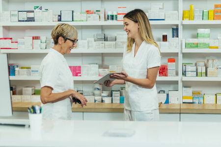 Senior female pharmacist using a smartphone and a tablet while working with her young colleague in the pharmacyの写真素材
