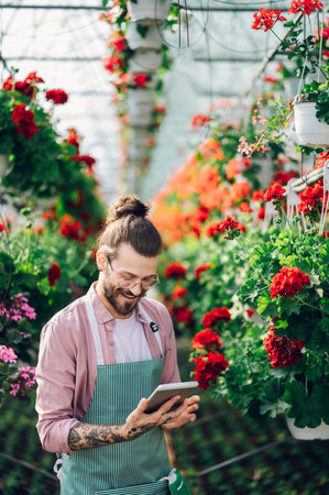 Cheerful young caucasian man checking information about flower delivery order on a tablet computer when working at a plant nursery. Young bearded smiling nursery garden worker wearing an apron.の写真素材