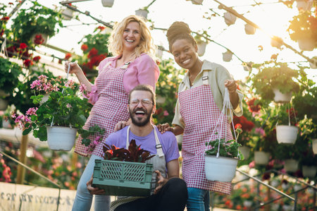 Garden center workers standing in greenhouse and working while holding flowers and smiling. Multiracial group of gardeners working and arranging colorful flowers in a sunny industrial plant nursery.の写真素材