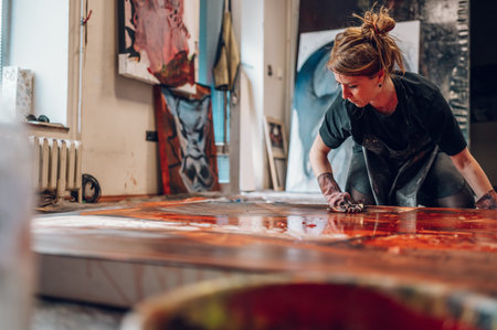 Portrait of a gorgeous redhead female artist working on several art projects on her bright studio. Young caucasian artistic woman using painting tools on a large canvas while drawing picture in studio.の写真素材