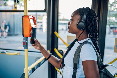 Young african american woman wearing earphones and paying for her bus ticket via smartphone. Black woman paying wireless for a ticket while in a bus during her morning commute. Payment and banking.の写真素材
