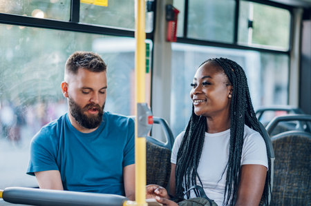 Young multiracial couple chatting on a bus together. Two diverse friends are talking while sitting in a bus. People in a public transport concept. Focus on a beautiful african american woman.の写真素材