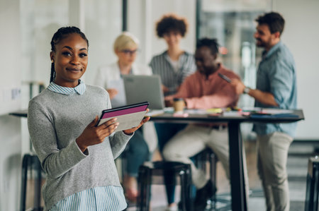 Positive black female executive standing in an office and using a tablet with colleagues working in background. African american business woman posing in the office and looking into the camera.の写真素材