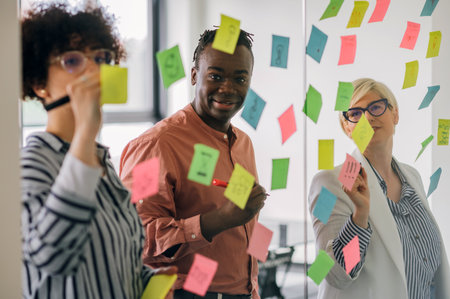 Focused young diverse teammates and their team leader working on an IT startup project and discussing details and tasks while writing notes on colorful paper stickers on a glass wall in the office.の写真素材