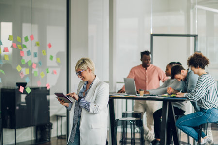Positive blonde female executive standing in an office and using a tablet with colleagues working in background. Smiling caucasian business woman posing in the office.の写真素材