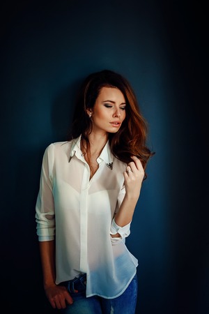 Beautiful girl standing near the dark blue wall in darkness in blue jeans and white shirtの写真素材