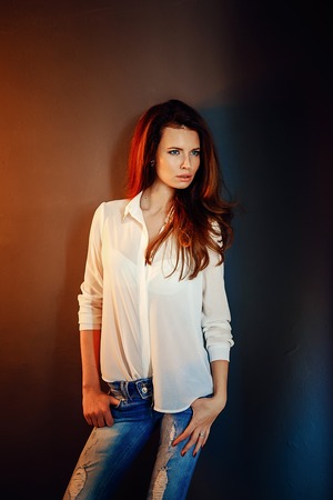 Beautiful girl standing near the dark blue wall in darkness in blue jeans and white shirtの写真素材