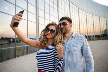 Young happy fashion couple in casual clothing with dark sungklasses taking selfie with a smart phone near the glass windows of the shop or cafe, summertime vacationの写真素材