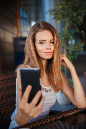 Awesome beauty caucasian girl in the white shirt sitting in the cafe looking at the phone, tabletの写真素材