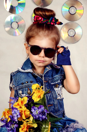 Little girl of 3 or 4 years old, light brown hair, dressed in blue jeans, blue gloves, dark glasses, holding a bouquet of bright flowers and CD's on backgroundの写真素材
