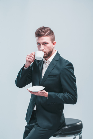 Studio shot of a man with white coffee cup, isolated on gray, sitting on the bar stoolの写真素材