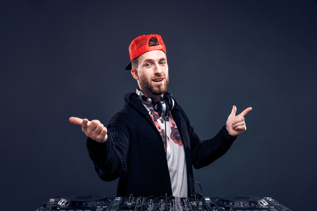 Closeup portrait of confident DJ with stylish red cap and headphones on neck mixing music on mixer looking up while standing isolated on dark backgroundの写真素材