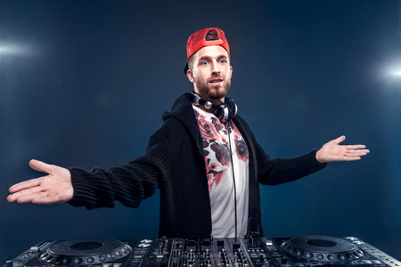 Closeup portrait of confident DJ with stylish red cap and headphones on neck mixing music on mixer looking up while standing isolated on dark backgroundの写真素材