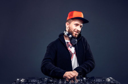 Closeup portrait of confident DJ with stylish red cap and headphones on neck mixing music on mixer looking up while standing isolated on dark backgroundの写真素材